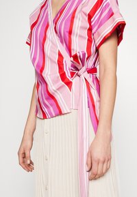 Haut à rayures roses, rouges et violettes, avec des manches courtes et une taille nouée. Le matériau semble léger avec une texture lisse.