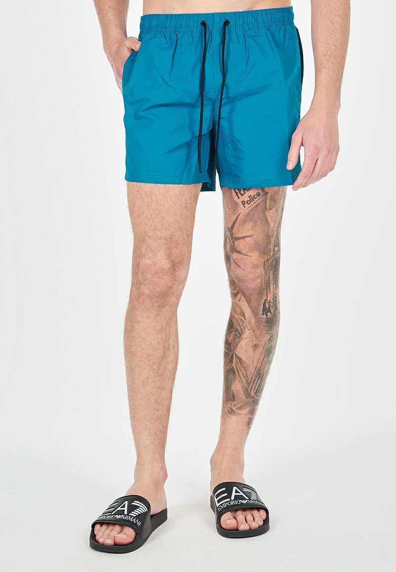 Pantaloni da bagno turchesi con vita elastica e cordino nero. Indossati con sandali neri con fascia e logo bianco.