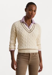 Sweter w kolorze kremowym, dziany, z wzorem w warkocze oraz dekoltem w serek. Nałożony na jasnofioletową koszulę z kołnierzykiem. Model ma na sobie brązowe spodnie z korduroy.