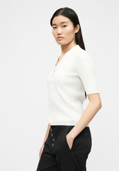 Νεαρή γυναίκα με μακριά σκούρα μαλλιά φοράει λευκό κοντομάνικο μπλουζάκι με γιακά και μαύρο παντελόνι με διακοσμητικά κουμπιά, ποζάροντας με το ένα χέρι στην τσέπη.