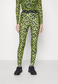 Czarne legginsy z jasnym zielonym wzorem serc, elastyczny materiał i czarny elastyczny pas z napisem. W zestawie z czarnymi butami sportowymi.