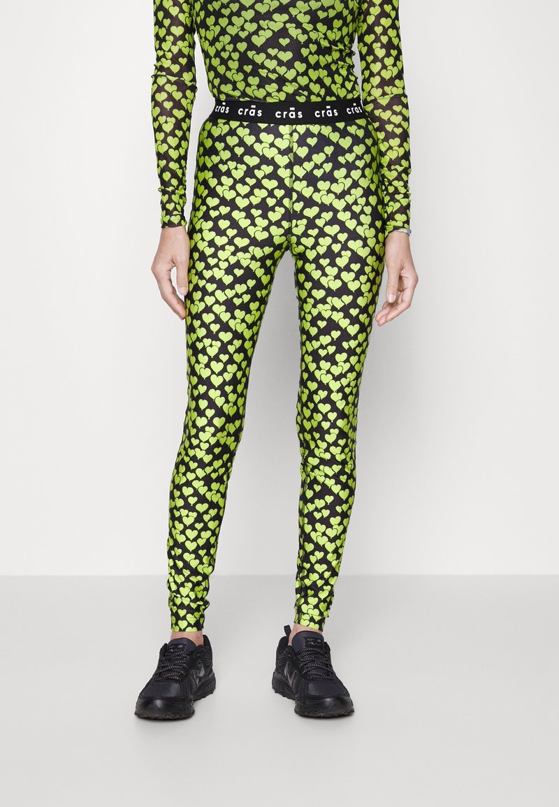 Czarne legginsy z jasnym zielonym wzorem serc, elastyczny materiał i czarny elastyczny pas z napisem. W zestawie z czarnymi butami sportowymi.