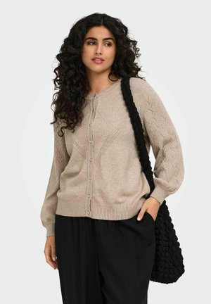 Femme aux cheveux bouclés portant un cardigan beige en tricot et un pantalon noir, portant un sac à épaule noir texturé, regardant sur le côté.