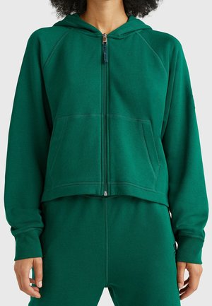 Personne portant un sweat à capuche zippé vert foncé et un pantalon assorti, debout les bras détendus le long du corps devant un fond uni.