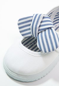 Zapatilla de lienzo blanca con puntera redondeada, tira de tela elástica y un lazo de rayas azules y blancas. Suela texturizada para agarre.