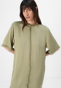 Olivegrünes, übergroßes T-Shirt mit kurzen Ärmeln, das Fransenverzierungen entlang der Kanten und eine zentrale Naht aufweist. Glatte Textur.