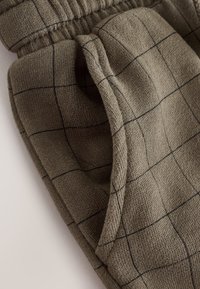 Brown knit pants with black grid pattern and elastic waistband, showing side pocket and textured fabric detail.