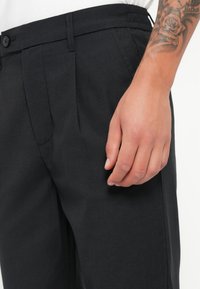 Schwarze, strukturierte Hosen mit Falten, einem Knopfverschluss und einem Bund. Eine Hand ruht auf dem Oberschenkelbereich zur Verdeutlichung.
