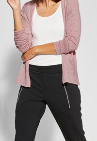 Hellrosa gestrickte Strickjacke über einem weißen Oberteil, kombiniert mit schwarzen Hosen mit weißen Seitenstreifen. Weiche Texturen und lässiges Design.