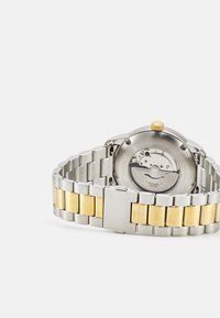 Montre en acier inoxydable argenté avec un bracelet aux accents dorés. Elle possède un boîtier rond, une couronne jaune et un fond transparent révélant les détails du mouvement.