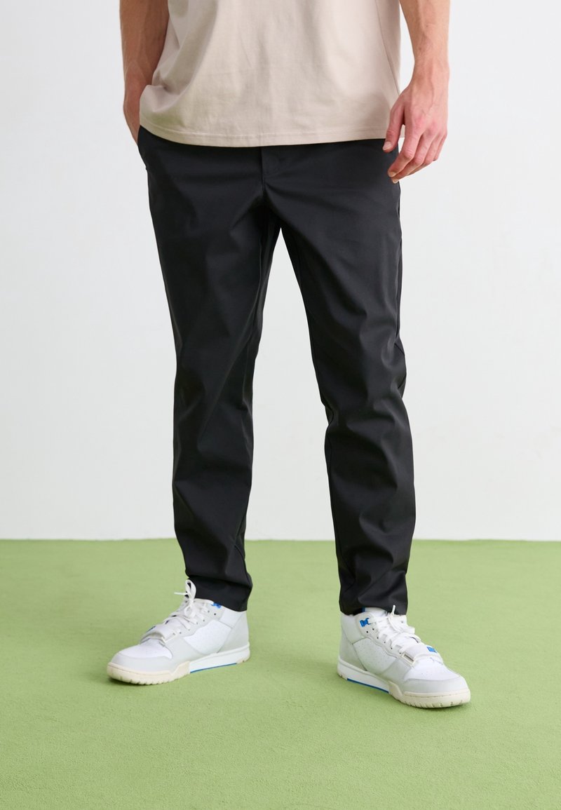 Homme portant un pantalon noir slim et des baskets blanches, debout sur un sol vert clair avec un fond blanc.