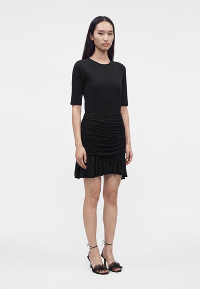 Femme debout devant un fond uni, portant un haut noir ajusté à manches courtes, une mini-jupe noire froncée avec ourlet à volants et des sandales noires.