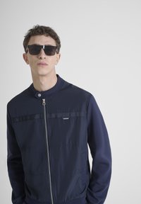 Navy plavi bomber jakna s kontrastnom teksturom, koja ima patentni zatvarač sprijeda, stojeći ovratnik i mali logotip na prsima.