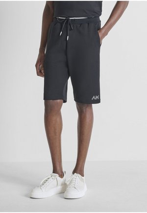 Pantaloncini neri con coulisse indossati da una persona con le mani nelle tasche, abbinati a scarpe da ginnastica bianche su uno sfondo chiaro uniforme.