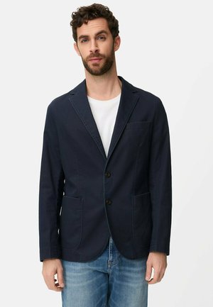 Mann trägt einen dunkelblauen Blazer über einem weißen T-Shirt und hellblauen Jeans, steht vor einem einfarbigen weißen Hintergrund.