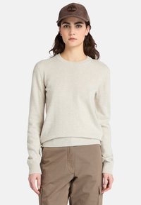 Giovane donna che indossa un maglione beige, pantaloni cargo marroni e un berretto in velluto a coste marrone con un logo a forma di albero, in piedi davanti a uno sfondo semplice.