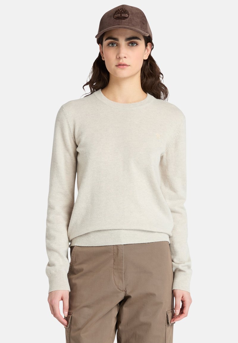 Giovane donna che indossa un maglione beige, pantaloni cargo marroni e un berretto in velluto a coste marrone con un logo a forma di albero, in piedi davanti a uno sfondo semplice.