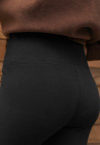 Nærbillede af en person iført sorte højtaljede leggings og en brun tekstureret top foran en træbaggrund.