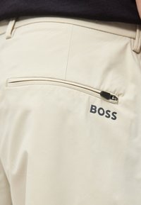 Μπεζ παντελόνι με φερμουάρ στην πίσω τσέπη και τη λέξη «BOSS» τυπωμένη με μαύρα γράμματα κοντά στην τσέπη στη δεξιά πλευρά.