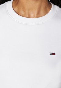White sweatshirt with a crew neck and ribbed texture. Features a small embroidered logo in red, blue, and white at the chest.