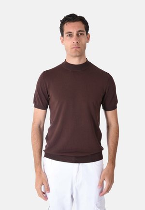 Uomo con capelli corti e scuri che indossa una maglia marrone a maniche corte e pantaloni bianchi, in piedi davanti a uno sfondo chiaro e semplice.