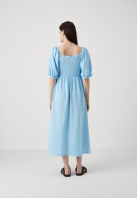 Rochie bleu deschis, până la genunchi, cu mâneci bufante și decolteu pătrat. Prezintă un spate plisat și un material texturat. Este purtată cu sandale închise la culoare.