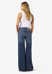 Žena s dlouhými blond vlasy, která má na sobě bílé tílko bez rukávů a modré džíny s širokými nohavicemi a vysokým pasem, stojící zády k nám.