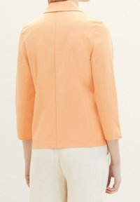 Veste couleur pêche avec un col classique, des manches trois-quarts et une texture lisse. Présente une couture droite dans le dos et un design minimaliste.