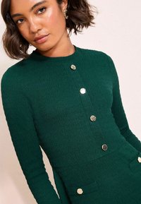 Groene jurk met textuur, lange mouwen, gouden knopen en twee zakken. De stof lijkt geribbeld, wat een subtiel patroon creëert.