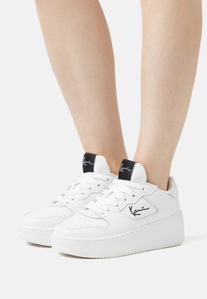 Białe skórzane trampki z grubą podeszwą, okrągłym noskiem, czarnymi akcentami na języku oraz charakterystycznymi detalami po bokach.