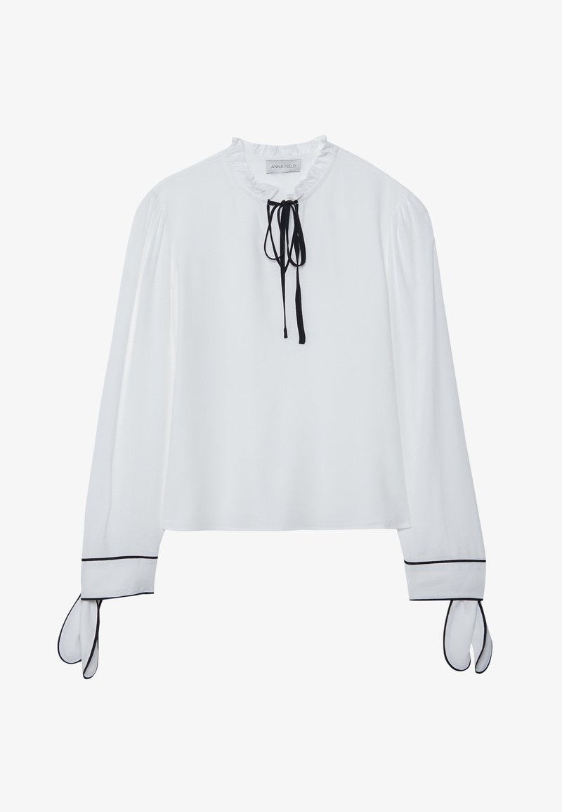 Bijela bluzica s ruflenim izrezom, crnom mašnom i dugim rukavima s crnim detaljima na manžetama. Glatka tkanina, opušteni kroj.