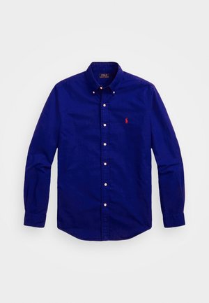 Chemise bleu roi à manches longues, boutonnée, avec col et petit logo rouge brodé sur la poitrine gauche.