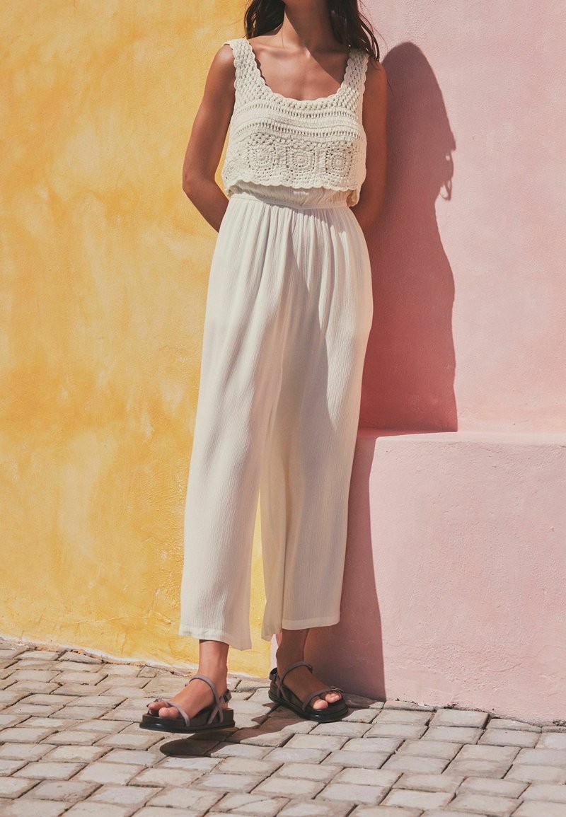 White, sleeveless top with textured crochet detailing paired with flowing white pants, complemented by black strappy sandals. Wall features warm yellow and pink tones.