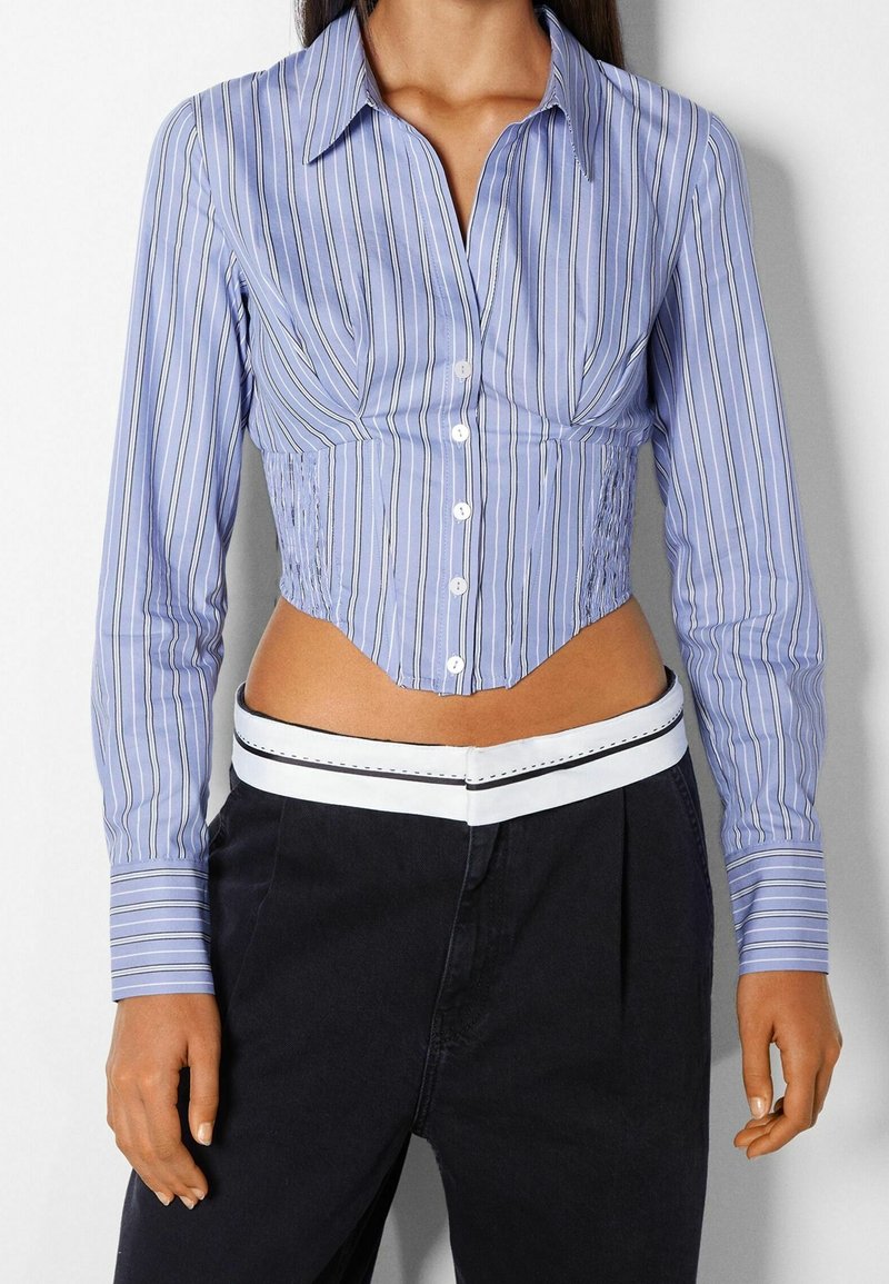 Blårandig kortärmad skjorta med skräddarsydd passform, knäppning framtill, skarp krage och texturerad sidofåll; ihopparad med mörka byxor med en vit accent vid midjan.