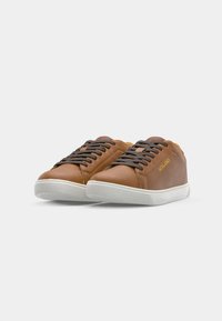 Bruine leren sneakers met witte rubber zolen, donkerbruine veters en een gouden logo aan de zijkant. Glad oppervlak met een ronde vorm.