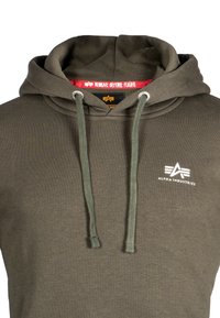 Felpa verde oliva con cappuccio, caratterizzata da un cordino, tasca anteriore e logo bianco. Etichetta interna con la scritta "RIMUOVERE PRIMA DEL VOLO" in rosso. Texture del tessuto morbida.