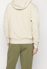 Osoba oblečená v světle béžovém mikině s tečkami a olivově zelených kalhotách, stojící zády k fotoaparátu před jednobarevným pozadím.