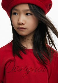 Rotes Langarmshirt mit "Desigual" aus Strasssteinen, kombiniert mit einer roten Filzberet. Dunkles Haar verdeckt teilweise das Shirt. Strukturierte Stoffdetails sind sichtbar.