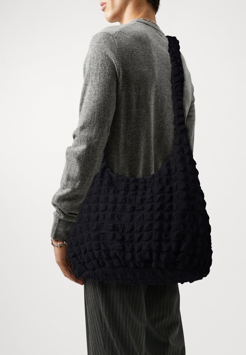 Personne portant un pull gris et un pantalon rayé avec un grand sac à épaule noir texturé sur un fond blanc uni.