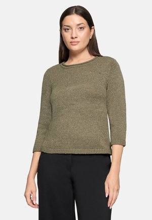 Kvinde iført en olivengrøn striktrøje med trekvartærmer og sorte bukser, stående mod en enkel lys baggrund.