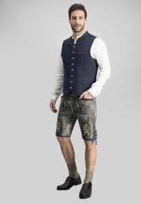 Traditionelle Kleidung mit einer blauen Wollweste, grauen Ledershorts mit gesticktem Löwendesign, grauen Socken und schwarzen Schuhen.