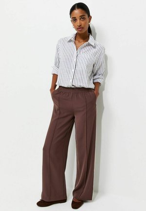 Femme debout, les mains dans les poches, portant une chemise blanche à rayures et un pantalon marron ample avec des ballerines marron.