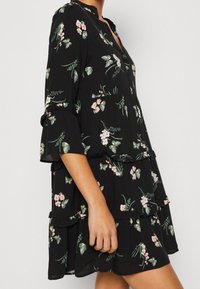 Schwarzes Kleid mit Blumenmuster, Rüschenschichten und 3/4-Ärmeln im Glockenschnitt. Hergestellt aus leichtem Stoff, mit einem bescheidenen Ausschnitt und leicht übergroßer Passform.