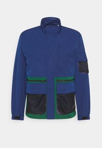 Blå jacka med hög krage, gröna och svarta nätfickor, framdragkedjor och lila detaljer. Tillverkad av hållbart tyg med en strukturerad design.