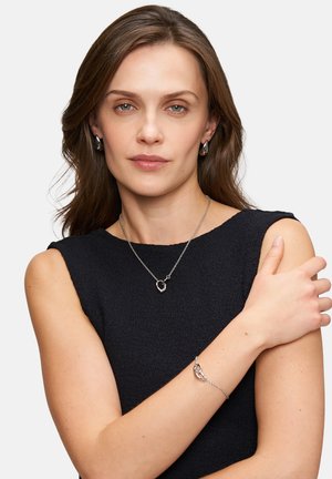 Zilveren kettingjuwelen omvatten een ketting met een harthanger en een armband. Het model draagt een zwart mouwloos topje.