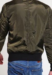 Muž v olivově zelené saténové bomber bundě a modrých džínách, zobrazený zezadu na jednobarevném pozadí.