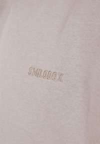 Beiger Stoff mit dem eingestickten "SMILODOX" in passendem Garn, das feine Strickstruktur und subtile Schatten zeigt.