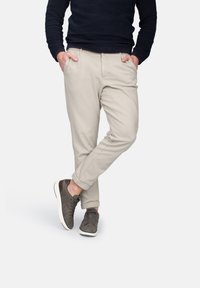 Pantaloni tapered beige con tasche laterali, abbinati a sneakers grigie. I pantaloni sono realizzati in un tessuto liscio, presentano una vestibilità comoda e orli arrotolati.