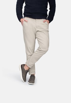 Pantalones tapered beige con bolsillos laterales, combinados con zapatillas grises. Los pantalones están hechos de una tela suave, presentan un corte relajado y puños enrollados.