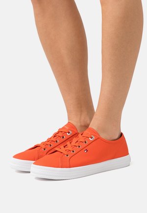 Personne portant des baskets basses en toile orange vif avec des semelles en caoutchouc blanches, des lacets blancs et un petit logo sur le côté.